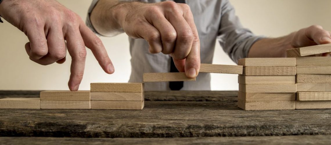 Businessman building staircase with wooden blocks to span a gap for partner to walk his fingers across towards promotion and progress.